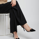 Person wearing black high-heeled shoes and black pants sitting on a white stool against a light gray background.