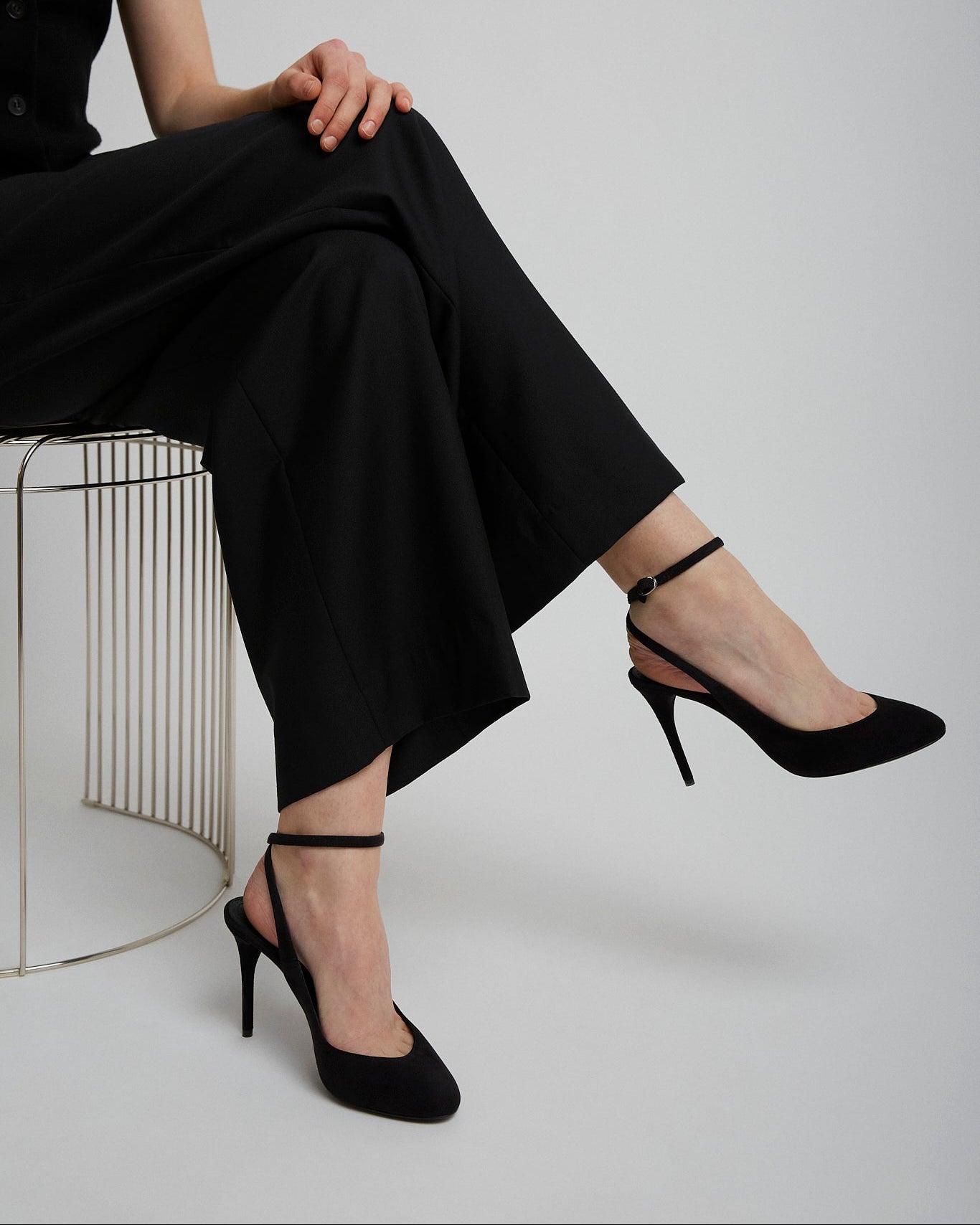 Person wearing black high-heeled shoes and pants sitting on a chair against a plain background