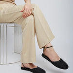 Person wearing beige pants and black shoes sitting on a wire stool against a white background