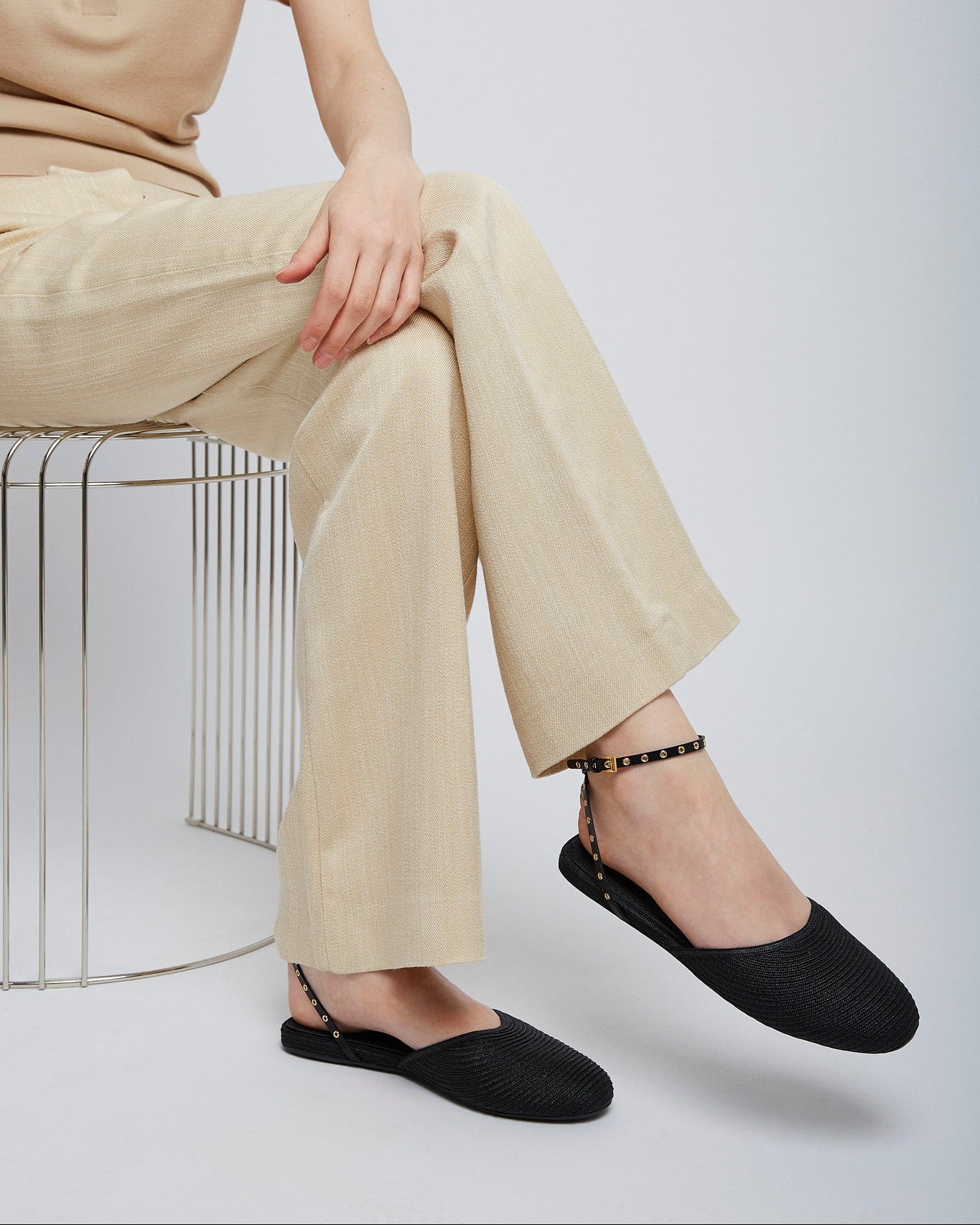 Person wearing beige pants and black shoes sitting on a wire stool against a white background