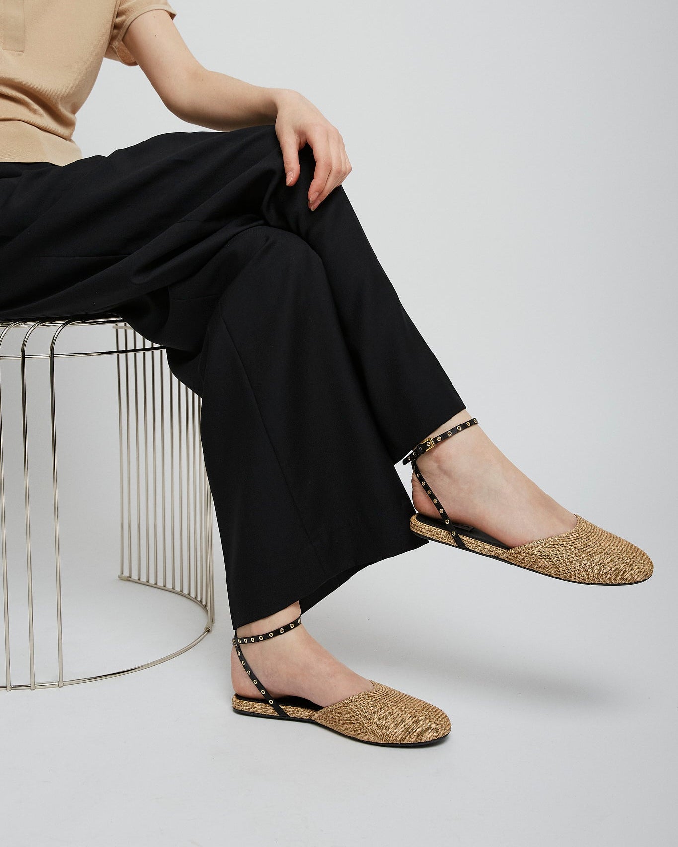Person wearing beige flats and black pants sitting on a wire stool against a white background