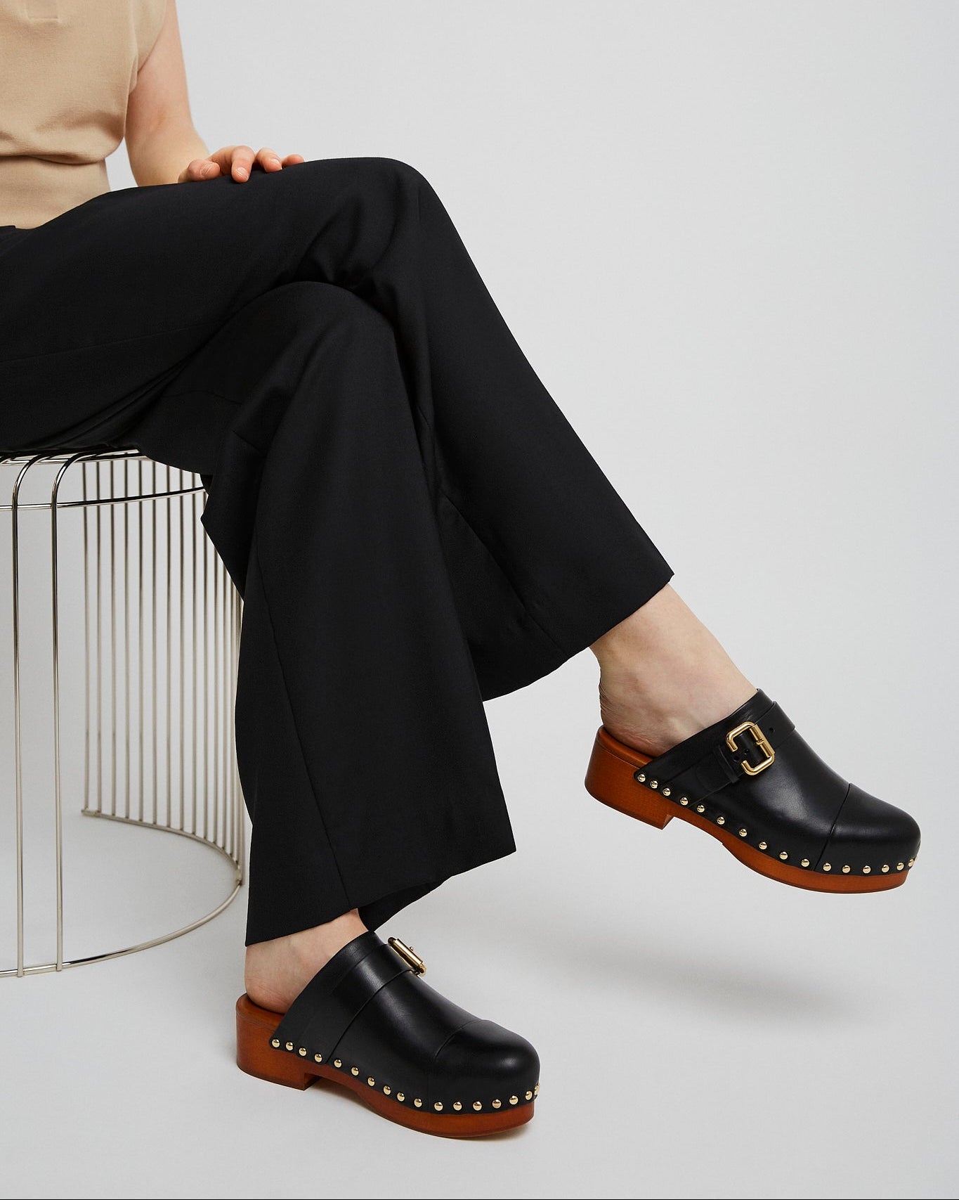 Person wearing black clogs with brown soles and a gold buckle, sitting on a wire chair.
