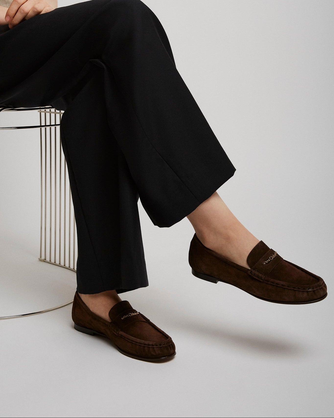 Person wearing dark brown loafers and black pants sitting on a white chair.