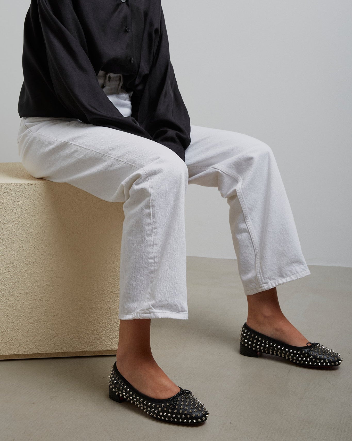 Person wearing a black shirt, white pants, and black studded flats sitting on a beige block.