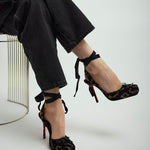 Person wearing black high-heeled shoes with ankle straps on a light gray background