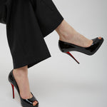 Black high-heeled shoes with red soles worn by a person in black pants on a light gray background