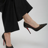 Black high-heeled shoes with red soles worn by a person in black pants on a light gray background