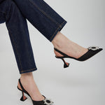 Black high-heeled shoes with decorative buckles worn with dark jeans on a light gray background