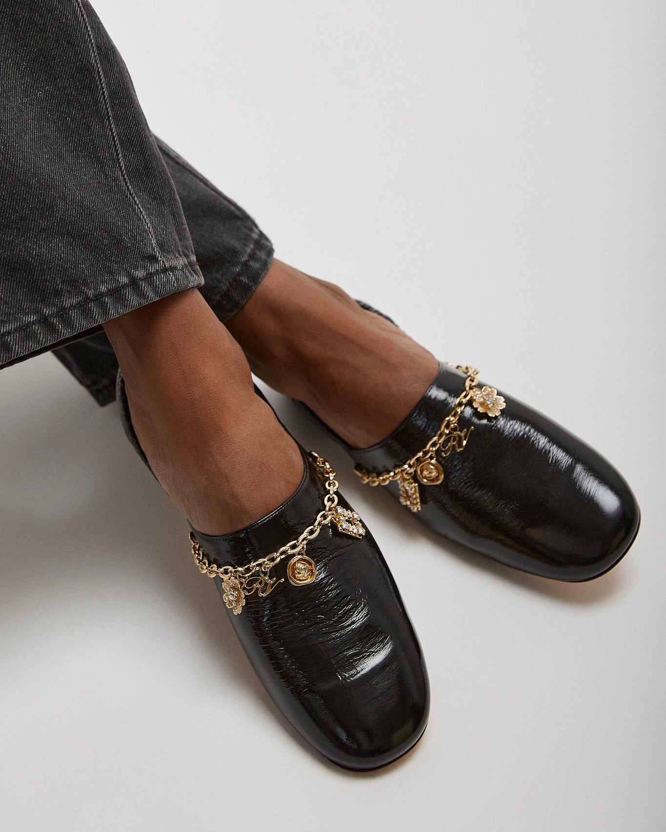 Black loafers with gold chain details worn with gray jeans on a light background