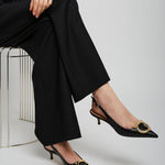 Person wearing black high-waisted pants and black high-heeled shoes with gold buckles on a white background