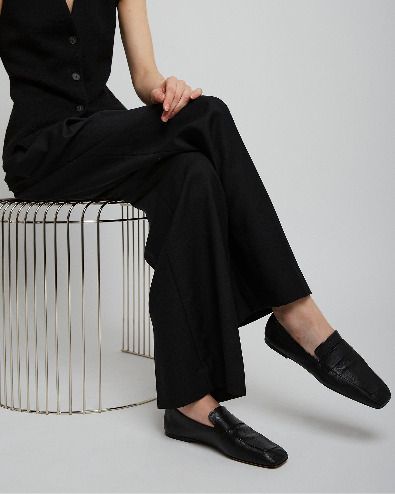 Person wearing a black sleeveless jumpsuit sitting on a metallic stool against a plain background