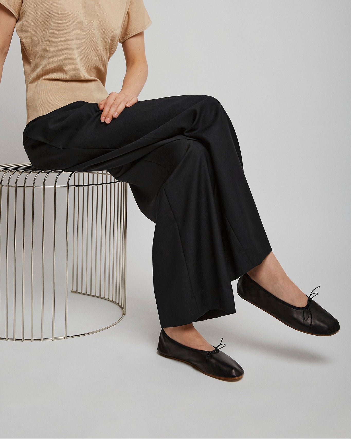 Person wearing a beige shirt, black pants, and black shoes sitting on a wire stool against a plain background.