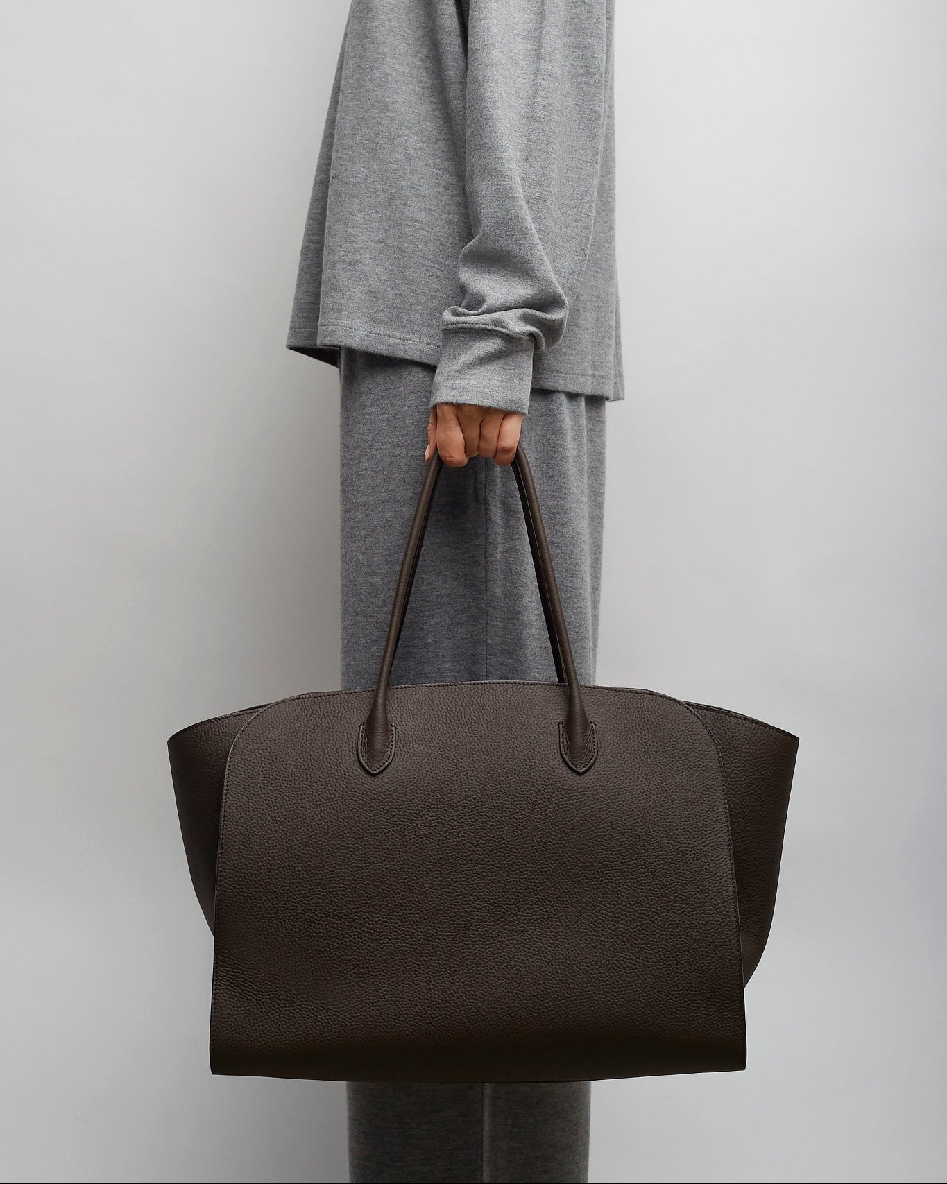 Person holding a dark brown leather tote bag against a plain background