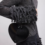 Person wearing a dark gray outfit with ruffled sleeves and holding a black heart-shaped clutch.