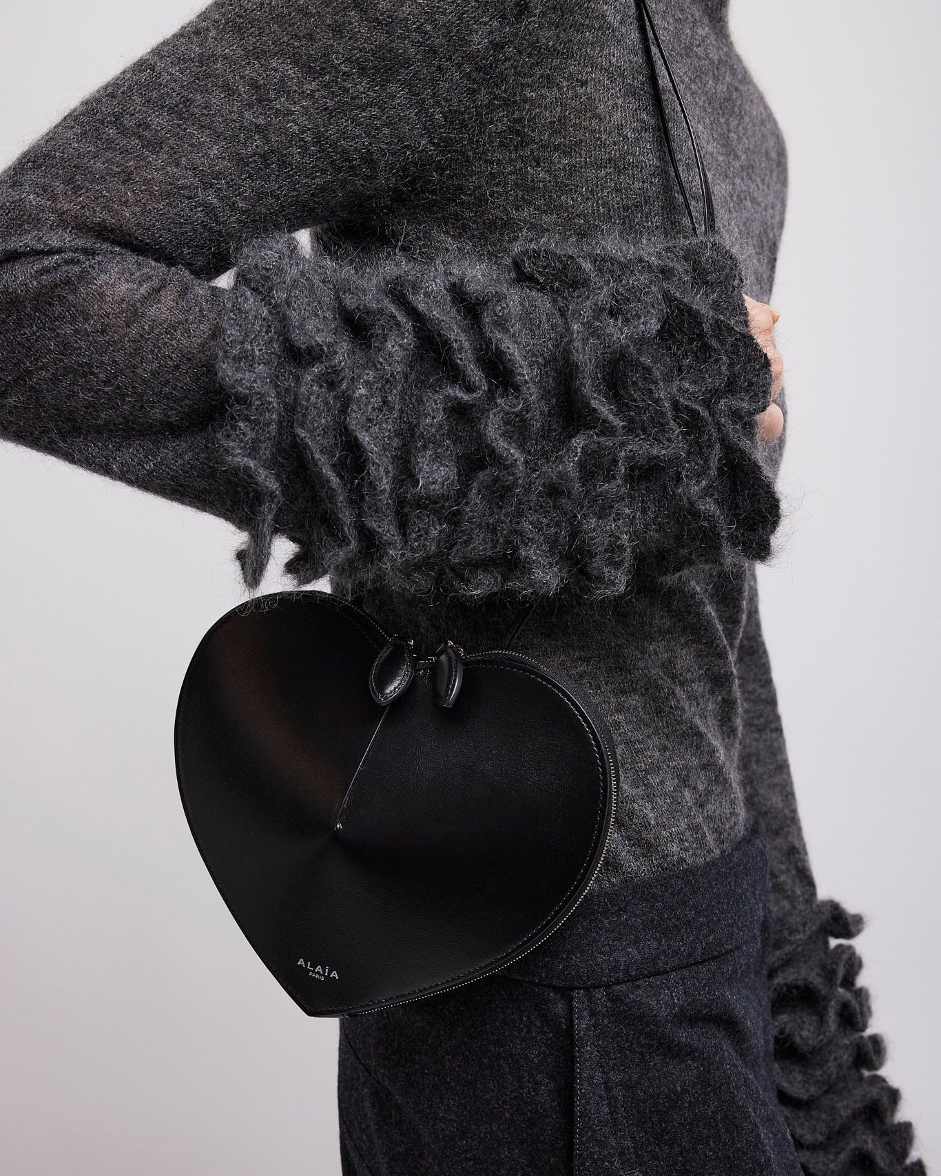 Person wearing a dark gray outfit with ruffled sleeves and holding a black heart-shaped clutch.