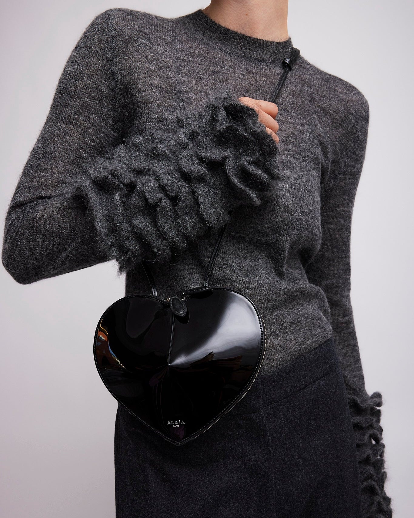 Person wearing a gray sweater with ruffled sleeves holding a black heart-shaped bag.