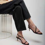 Person wearing black high-heeled sandals sitting on a wire stool against a plain background