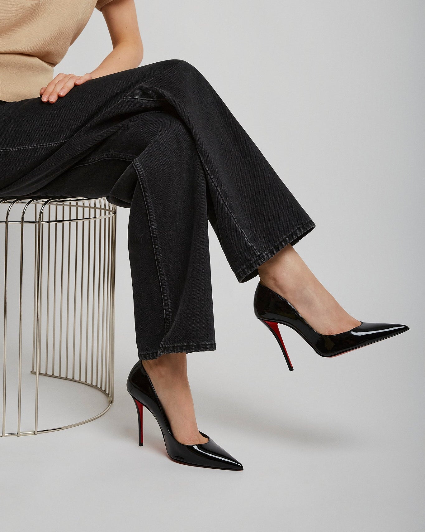 Person wearing black high-heeled shoes with red soles, sitting on a wire stool against a plain background.