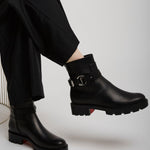 Black leather boots with a brand logo worn by a person sitting on a white chair.