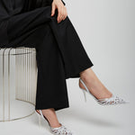 Person wearing a black outfit with white high-heeled shoes sitting on a metal stool against a white background