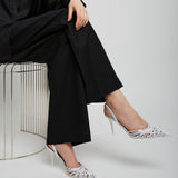 Person wearing a black outfit with white high-heeled shoes sitting on a metal stool against a white background