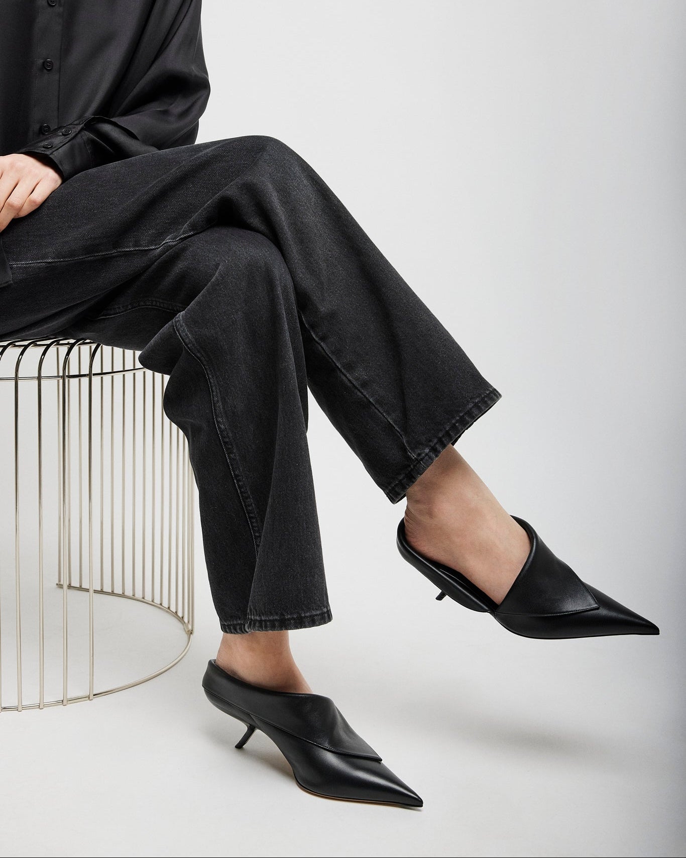 Person wearing black outfit with high-waisted pants and pointed-toe shoes sitting on a wire stool.